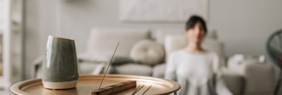 Persona meditando en casa sentada en el suelo, con incienso encendido sobre una mesa en primer plano, en un ambiente tranquilo y minimalista, durante su meditación online.
